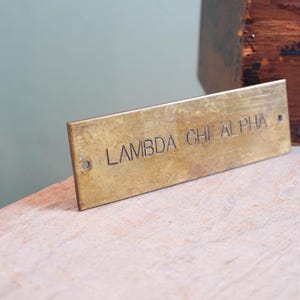 Pode incluir: Placa retangular de lat&atilde;o com o texto gravado "LAMBDA CHI ALPHA". A placa tem dois pequenos orif&iacute;cios em cada extremidade. A placa est&aacute; apoiada numa superf&iacute;cie de madeira clara, com uma caixa de madeira ao fundo.