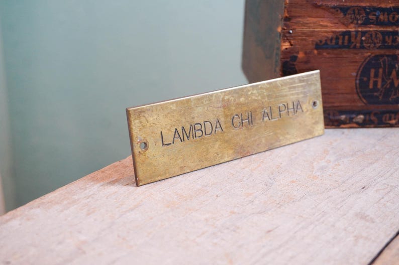 Pode incluir: Placa retangular de lat&atilde;o com o texto gravado "LAMBDA CHI ALPHA". A placa tem dois pequenos orif&iacute;cios em cada extremidade. A placa est&aacute; apoiada numa superf&iacute;cie de madeira clara, com uma caixa de madeira ao fundo.