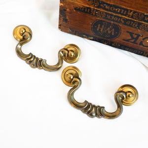 May include: Two antique brass drawer pulls with a decorative, curved design. Each pull features a circular backplate and a gracefully arched handle. The brass has a warm, aged patina, suggesting vintage charm. A wooden box with text is in the background.