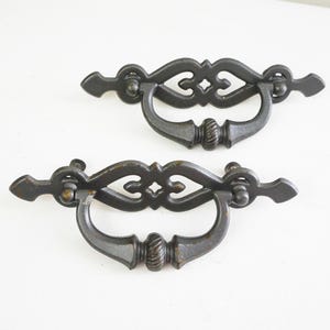 May include: Two antique-style, dark metal drawer pulls. Each pull features a curved handle with decorative detailing, including a central flourish and a ribbed section. The pulls have pointed end pieces and are designed for furniture.
