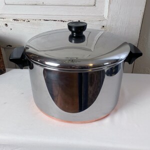 May include: A stainless steel pot with a lid and black handles. The pot has a copper-colored bottom and a black knob on the lid. The pot is sitting on a white surface.