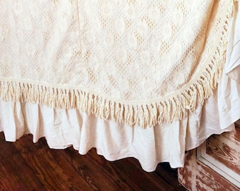 May include: A white crocheted throw blanket with a fringe border and a ruffled edge. The blanket is draped over a wooden surface.
