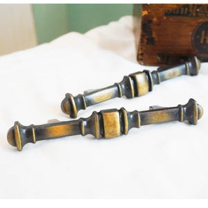 May include: Two antique bronze cabinet pulls with a detailed design. The pulls have a rectangular center section and rounded ends. The finish has a warm, aged appearance, with hints of gold and darker bronze tones. They are set against a white background.