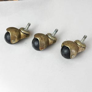 May include: Three ball casters featuring dark black spherical wheels housed in aged brass-toned metal sockets. Each caster includes a threaded metal stem with a washer at the base, designed for attachment to furniture legs for mobility and support.