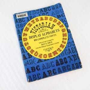 May include: A vintage book titled "Victorian Display Alphabets" with 100 complete fonts. The book has a blue cover with repeating "ABC" patterns and a yellow circular label. The label features the title and author's name.