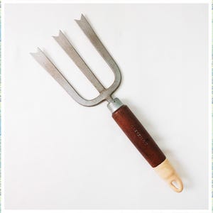 May include: A three-pronged garden tool with a brown wooden handle and a white plastic tip. The tool is made of metal and has a slightly curved shape.