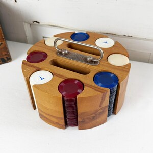 May include: A circular wooden poker chip carousel featuring a central metal handle and eight vertical slots. The carousel holds stacks of translucent red, blue, and white poker chips, with some white chips marked with a blue letter T on the top surface.