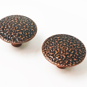 May include: Two bronze-colored cabinet knobs with a hammered texture. The knobs have a rounded, disc-like shape and a short stem. The finish is a dark, mottled bronze, and they are set against a white background.