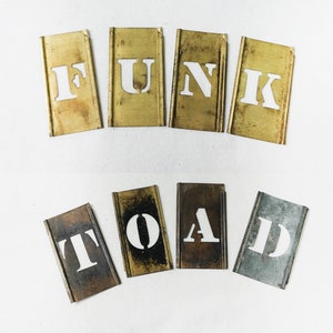 May include: A set of metal letter stencils spelling out the words "FUNK TOAD". The stencils are made of brass and have a worn, vintage look.