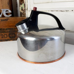 May include: A stainless steel kettle with a black handle and spout. The kettle has a copper-colored base and a red knob on the lid. The kettle is sitting on a white surface.