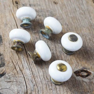 May include: Six white ceramic cabinet knobs with brass and aged metal bases are arranged on a weathered wooden surface. The knobs have a rounded shape with a flat top and a brass center.