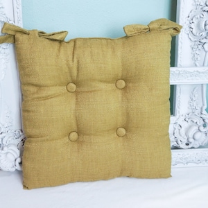 May include: A mustard yellow square cushion with four buttons. The cushion is made of a textured fabric and has a simple design.