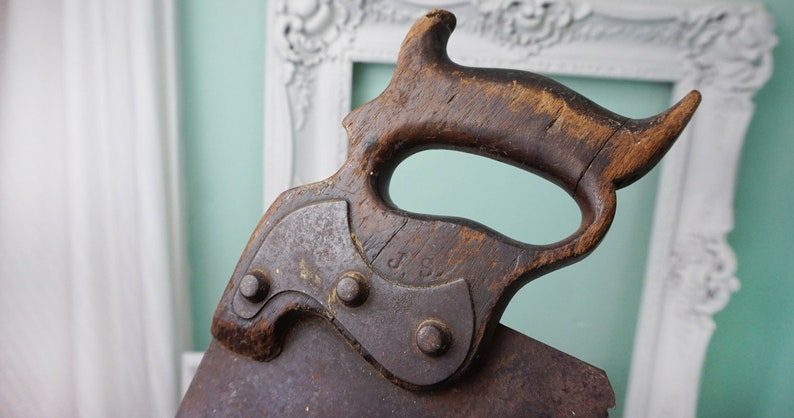 May include: Close-up of an antique hand saw with a wooden handle. The handle is dark brown and has a unique shape. The metal blade is rusty and has a J.S. marking.