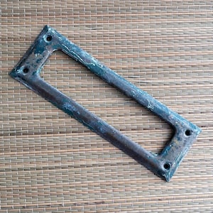 May include: Rectangular metal frame with a distressed, aged appearance. The frame has a dark, weathered finish with hints of blue-green patina. Four holes are visible at the corners, suggesting it may be a decorative or functional element.