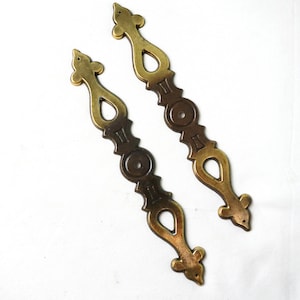 May include: Two ornate brass drawer pulls with a decorative design. The pulls are a dark brass color with a shiny finish.