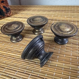 May include: Four antique brass cabinet knobs with a distressed finish. The knobs have a round, tiered design and are set against a natural woven background. One knob is lying on its side, showing its mounting hardware.