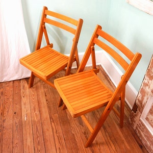 May include: Two wooden folding chairs with slatted seats and backs. The chairs are light brown in color and are sitting on a wooden floor.
