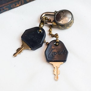 May include: Vintage brass padlock with two keys. The keys have black key fobs with the word "Lark" in cursive. The padlock is connected to the keys with a brass chain. The padlock and keys are on a white surface.