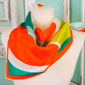 May include: A colorful silk scarf draped on a mannequin. The scarf features a vibrant design with orange, green, white, and yellow sections. The scarf is a square shape and appears to be made of a lightweight material.