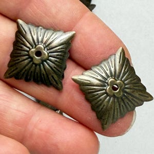 May include: Two square-shaped metal components with an antique brass finish. Each piece features a central four-petaled flower motif surrounded by radiating embossed lines that extend toward the pointed corners of the square frame.