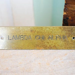 Pode incluir: Placa retangular de lat&atilde;o com o texto gravado "LAMBDA CHI ALPHA". A placa tem uma apar&ecirc;ncia desgastada e envelhecida com dois orif&iacute;cios para montagem. Uma caixa de madeira &eacute; vis&iacute;vel ao fundo.