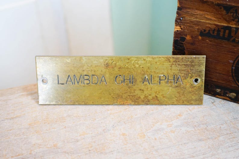 Pode incluir: Placa retangular de lat&atilde;o com o texto gravado "LAMBDA CHI ALPHA". A placa tem uma apar&ecirc;ncia desgastada e envelhecida com dois orif&iacute;cios para montagem. Uma caixa de madeira &eacute; vis&iacute;vel ao fundo.