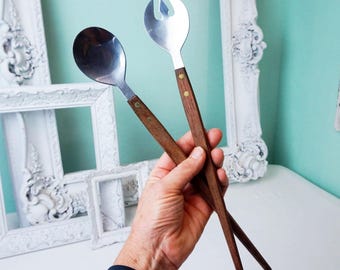May include: A pair of vintage salad servers. One server is a spoon, the other a fork. Both have stainless steel heads and dark wood handles, each secured with two brass rivets. The servers are held against a light blue background.