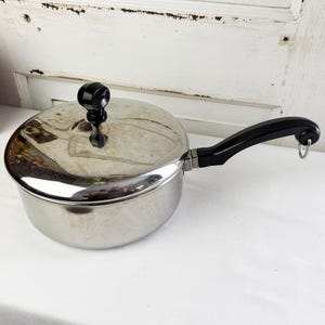 May include: A polished stainless steel saucepan with a matching lid, featuring a black heat-resistant handle and a black knob on the lid. The handle includes a small metal loop at the end for hanging, and the lid has a reflective metallic surface.