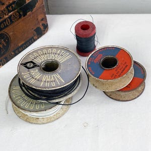 May include: A collection of vintage spools of wire and thread. The spools are made of metal and cardboard, with various colors and markings. One spool is red, and the others are blue, silver, and tan. The image also includes a wooden box.