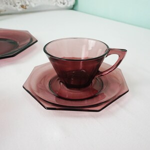 May include: A set of purple glass teacups and saucers with an octagonal shape. The cups and saucers are made of clear glass with a purple tint.