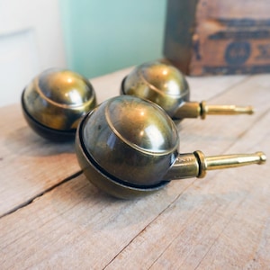 May include: Three antique brass casters with a spherical design and a cylindrical stem. The casters have a dark, aged patina, suggesting they are vintage. They are arranged on a light-colored wooden surface.