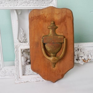 May include: A wooden plaque with a decorative brass door knocker in the shape of an urn. The knocker is attached to the plaque with two screws. The plaque has a distressed wood finish.