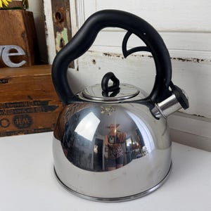 May include: A stainless steel whistling tea kettle with a polished, reflective finish. It features a black, curved, heat-resistant handle with an integrated trigger mechanism for the spout cover, and a matching black knob on the removable lid.