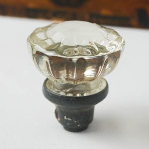 May include: A clear glass cabinet knob with a faceted, flower-like design. The knob sits atop a black base. The glass is slightly cloudy, and the knob appears to be vintage.