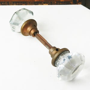 May include: Antique glass door knob with a faceted design. The knob has a clear, transparent appearance and is mounted on a brass-colored metal base and rod. The door knob is a vintage item.