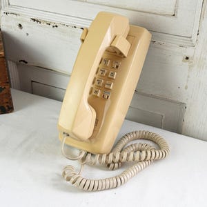 May include: A beige wall-mounted telephone featuring a rectangular base and a matching handset resting in the cradle. The device includes a standard numeric keypad with twelve buttons, each labeled with numbers and letters, and a coiled beige cord.