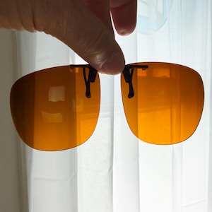 May include: A pair of amber-tinted clip-on sunglasses featuring two rounded lenses connected by a black metal bridge mechanism. The lenses have a warm orange hue and are designed to attach to existing eyewear frames with small black clips.