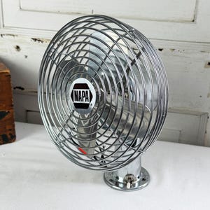 May include: A vintage-style chrome electric fan featuring a circular wire cage and a central emblem with the text NAPA. The fan has a metallic finish, three blades visible behind the protective grill, and a small toggle switch on the base.