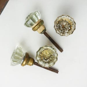 May include: Four vintage glass doorknobs with brass hardware and dark metal stems. Two knobs have a faceted, clear glass design, while the other two have a scalloped, textured appearance. The knobs are arranged on a white surface.