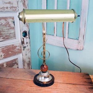 May include: A vintage brass desk lamp with a musical note design. The lamp features a long, rectangular shade and a base with a wooden sphere and a decorative silver and dark brown base. The lamp is on a wooden surface.