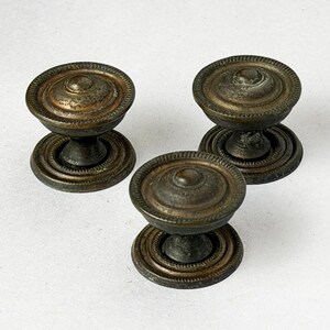 May include: Three circular metal knobs with a dark, weathered bronze finish. Each knob features a raised, rounded center dome surrounded by a textured, beaded edge and a matching circular base plate, showcasing a consistent aged metallic appearance.