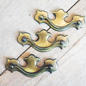 May include: Three antique brass drawer pulls with a decorative, curved design. Each pull features a central handle and ornate end pieces. The pulls are arranged on a light-colored wooden surface, showcasing their aged patina and vintage aesthetic.