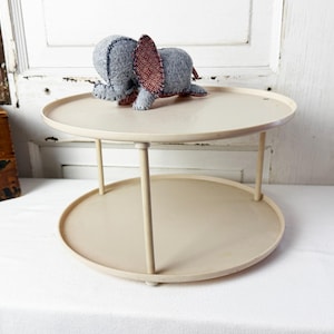May include: A two-tiered off-white circular stand with thin vertical supports. A small, handmade gray felt elephant with dark red patterned fabric ears sits on the top tier of the stand. The structure features two smooth, round trays.