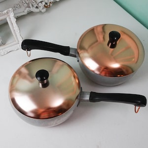 May include: Two copper-toned saucepans with black handles and lids. The saucepans are on a white surface.