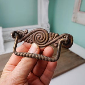 May include: A vintage brass drawer pull with a decorative scroll design and a twisted rope handle.