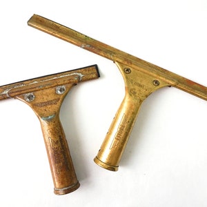 May include: Two vintage brass window squeegees with black rubber blades. The larger squeegee has the word "ETTORE" printed on the handle.