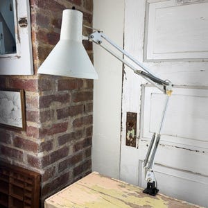 May include: A white metal architect lamp with an adjustable articulated arm and a conical shade. The lamp features a clamp base for attachment to a surface, exposed metal springs at the joints, and a simple white finish on the exterior components.