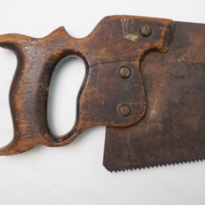 May include: A vintage hand saw with a wooden handle and a rusty metal blade. The handle has a unique shape with two finger holes and three screws.