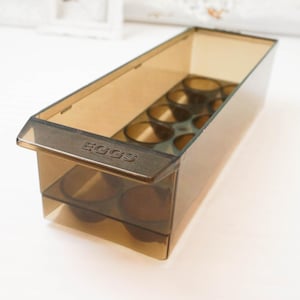 May include: A vintage brown plastic egg holder with the word "EGGS" embossed on the front. The container has individual compartments for holding eggs and is designed to fit in a refrigerator. The plastic is translucent, allowing visibility of the contents.