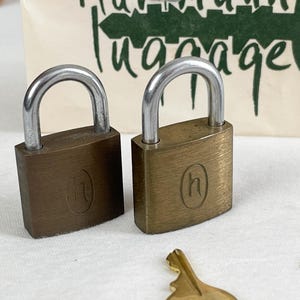 May include: Two vintage brass padlocks with silver-colored U-shaped shackles. Each lock has a rectangular body with a rounded top and the letter "h" engraved on the front. The background includes a cream-colored bag with the words "Hannah Luggage" in green.